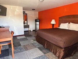 Habitación de hotel con cama, escritorio, silla, televisión, lámpara y kitchenette.