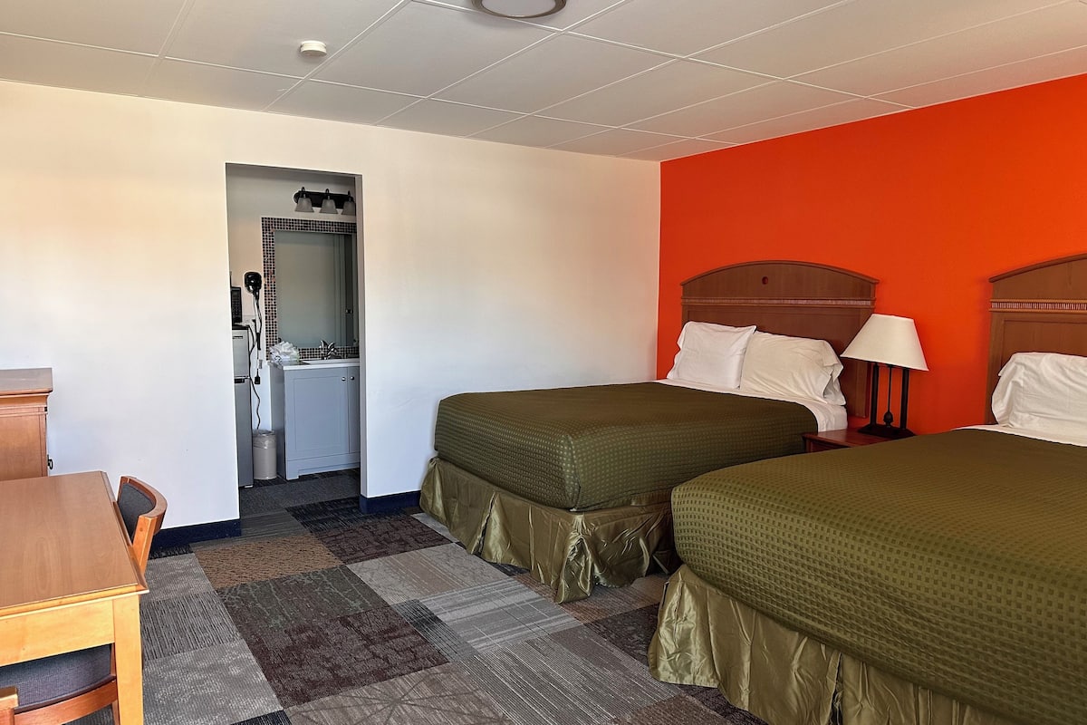 Habitación de hotel con dos camas individuales, un escritorio con silla, una mesita y un baño con lavamanos y espejo.