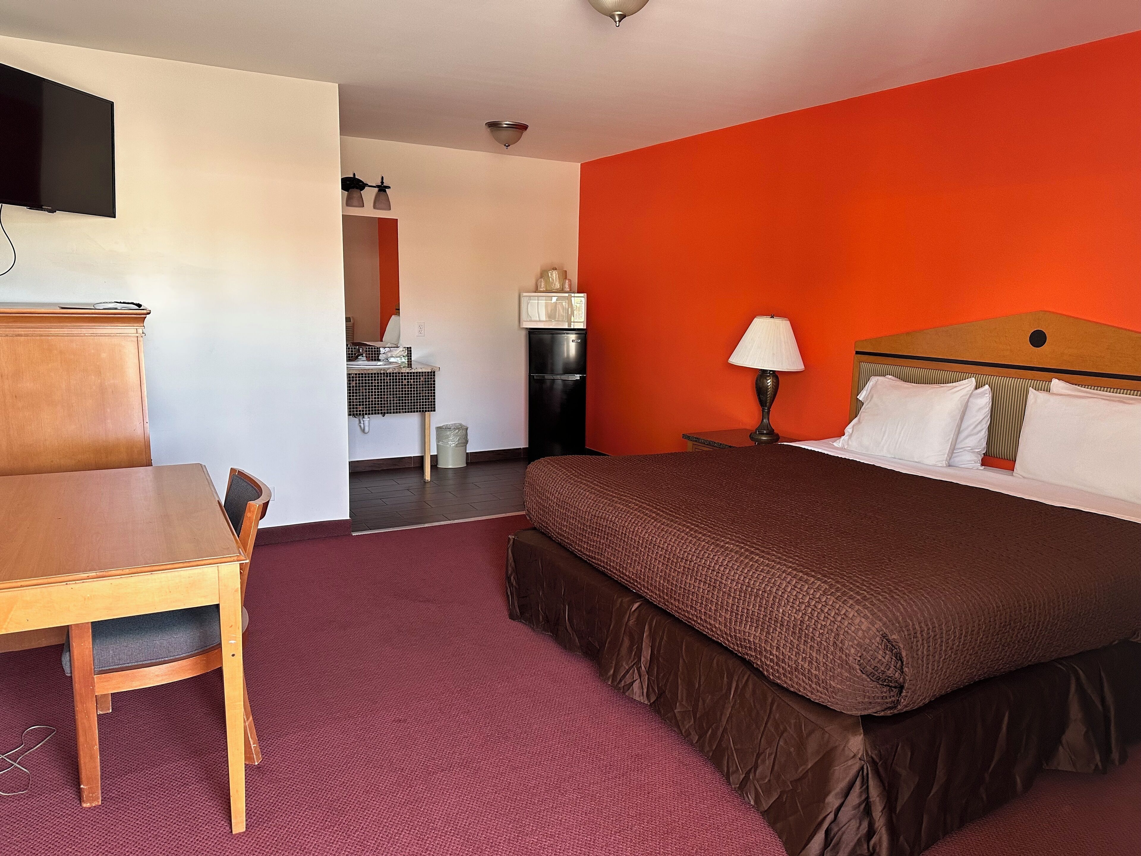 Ett hotellrum med en säng, ett skrivbord, en stol, en tv, en lampa och ett litet pentry.