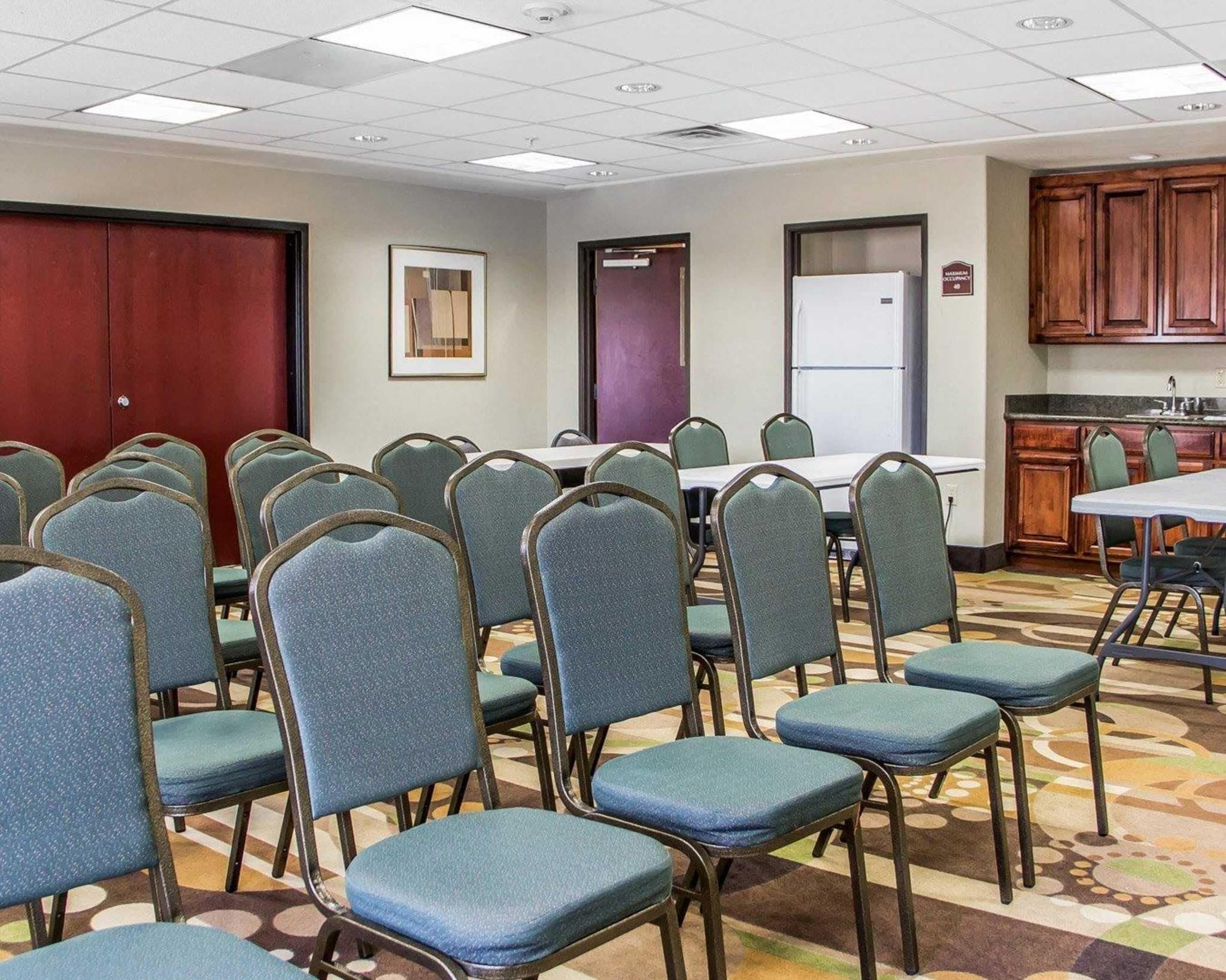 Et mødelokale med rækker af stole, et hvidt bord og et køkken med træskabe.