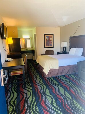 A hotel room with a bed, a desk with a lamp, a chair, a TV, and a painting on the wall.