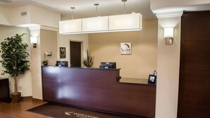 Een hotel receptie met een houten balie, een welkomstmat, een bloempot en een bord met de naam van het hotel.
