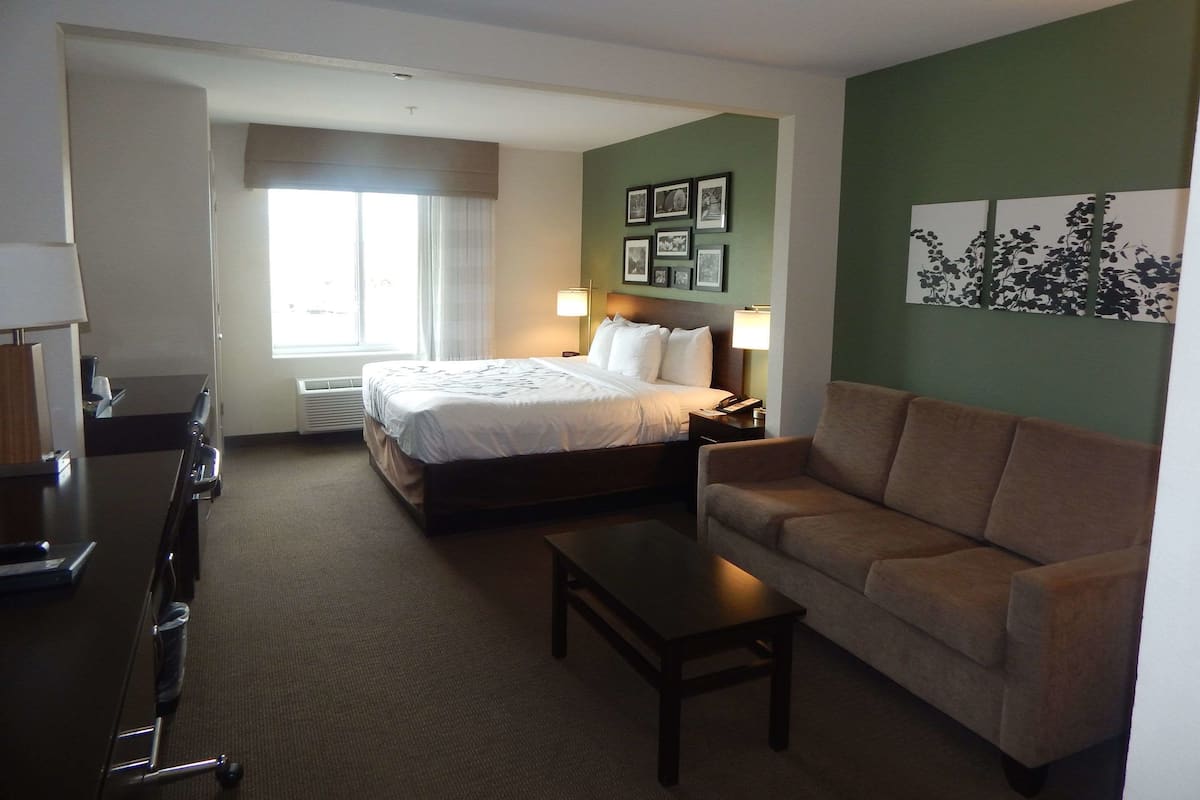 A hotel room with a bed, a sofa, a desk, and a chair. There is a window with curtains and a wall with framed pictures.