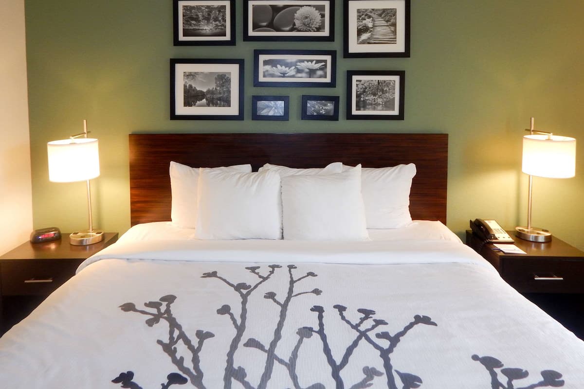 A hotel room with a neatly made bed, a wooden headboard, and a wall adorned with framed pictures. There are two bedside tables with lamps and a telephone on one.