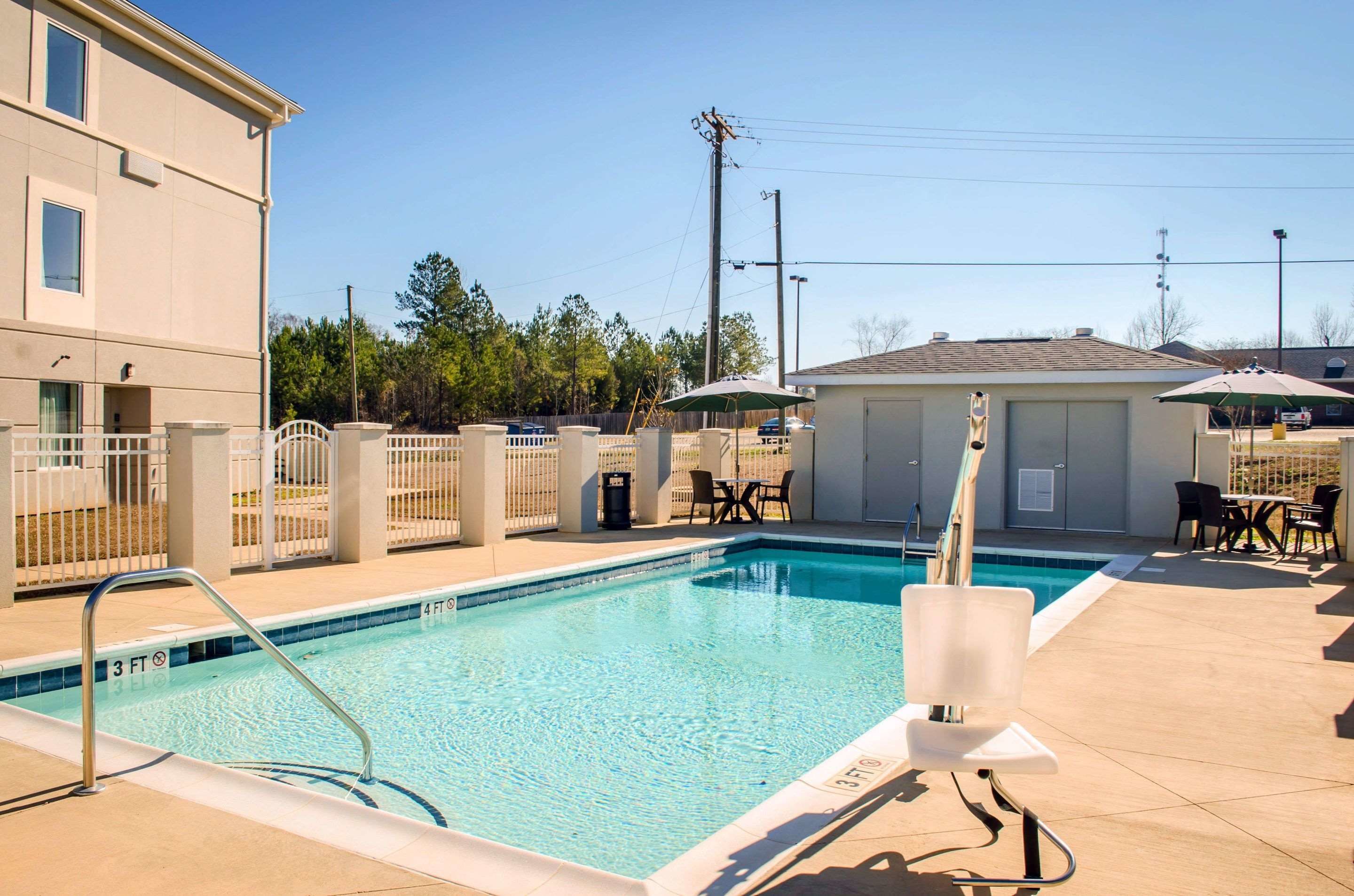 A rectangular swimming pool with a metal ladder, surrounded by a fence. There are outdoor tables and chairs, and a covered area with umbrellas.