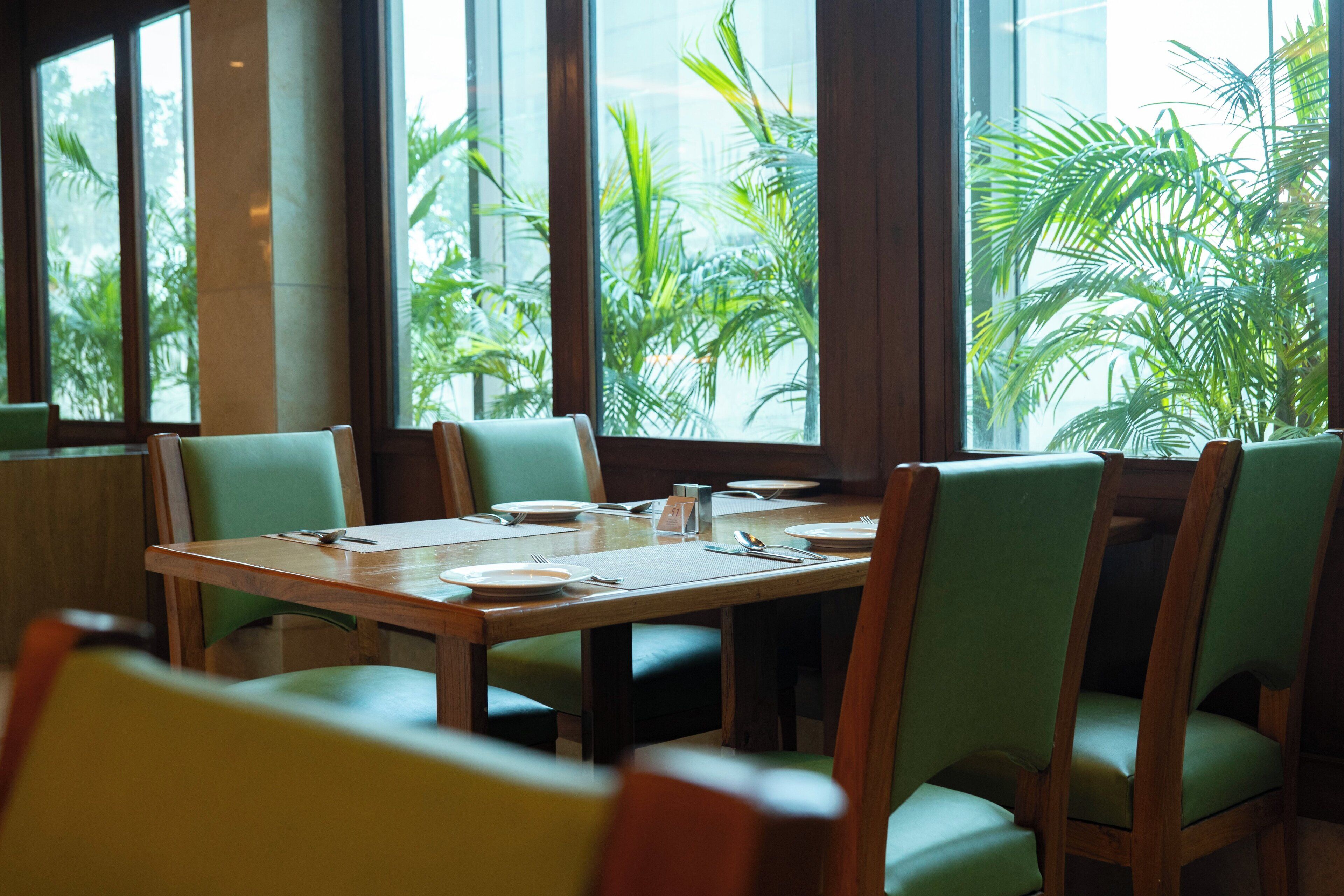 A dining area with a wooden table, green upholstered chairs, and large windows with a view of tropical plants.