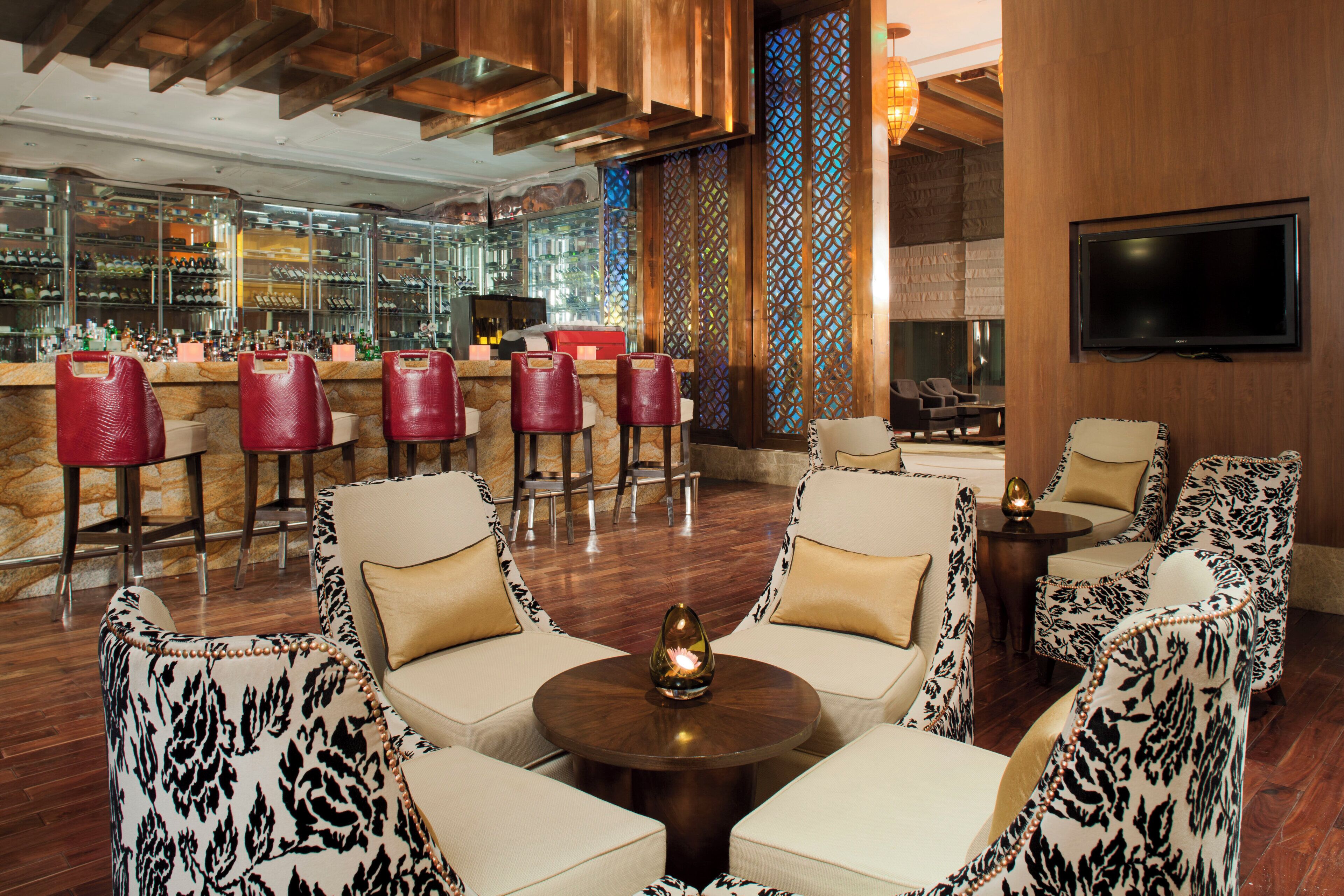A bar area with multiple red bar stools, a television mounted on the wall, and a seating area with patterned chairs and a round table.