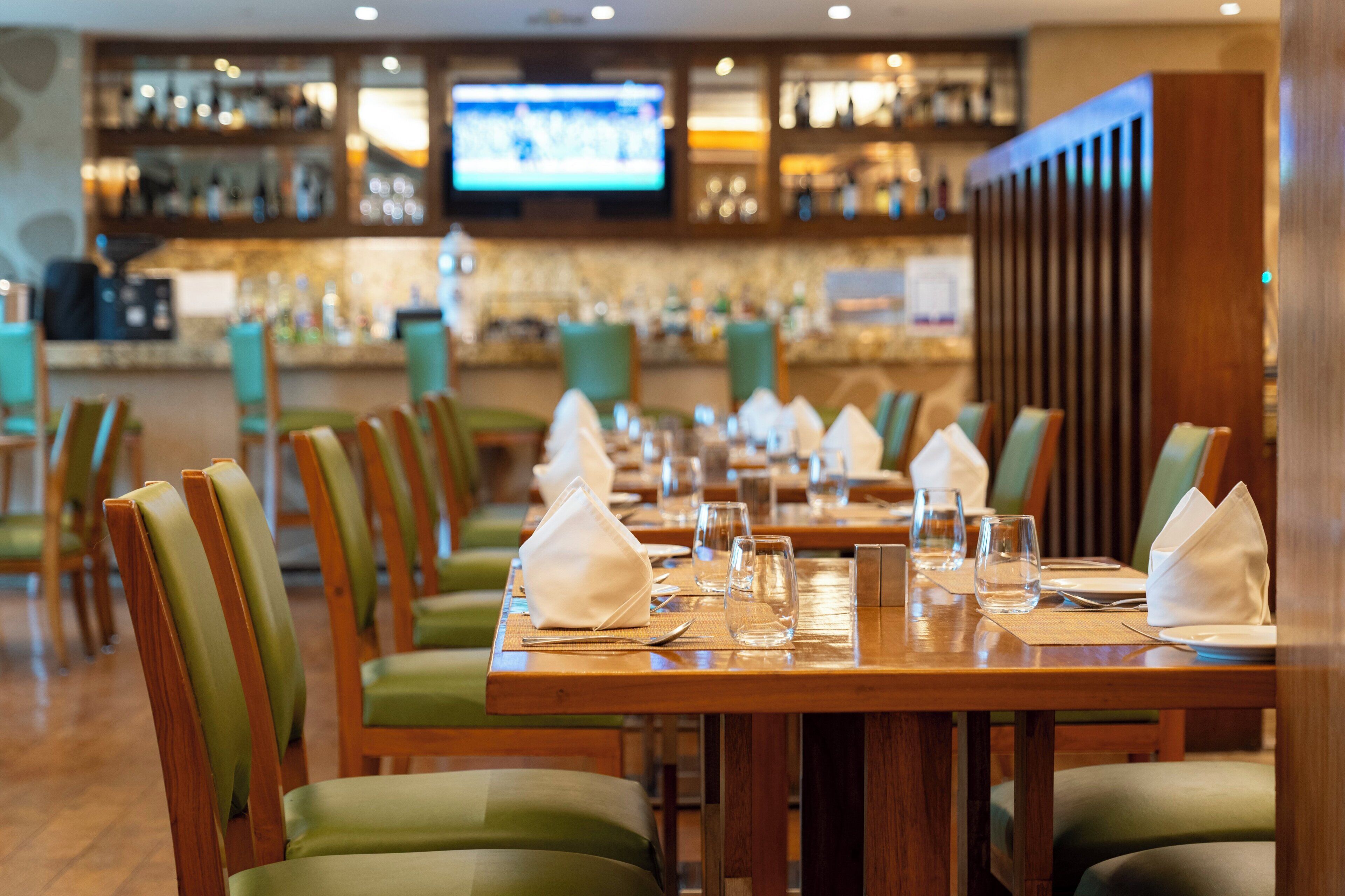 Un restaurante con mesas de madera y sillas verdes, una zona de bar con estantes llenos de botellas y una pantalla de televisión.