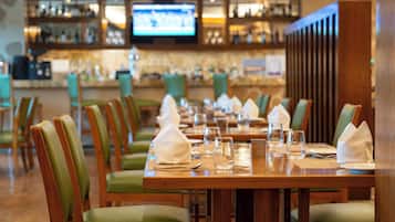 Un restaurante con mesas de madera y sillas verdes, una zona de bar con estantes llenos de botellas y una pantalla de televisión.