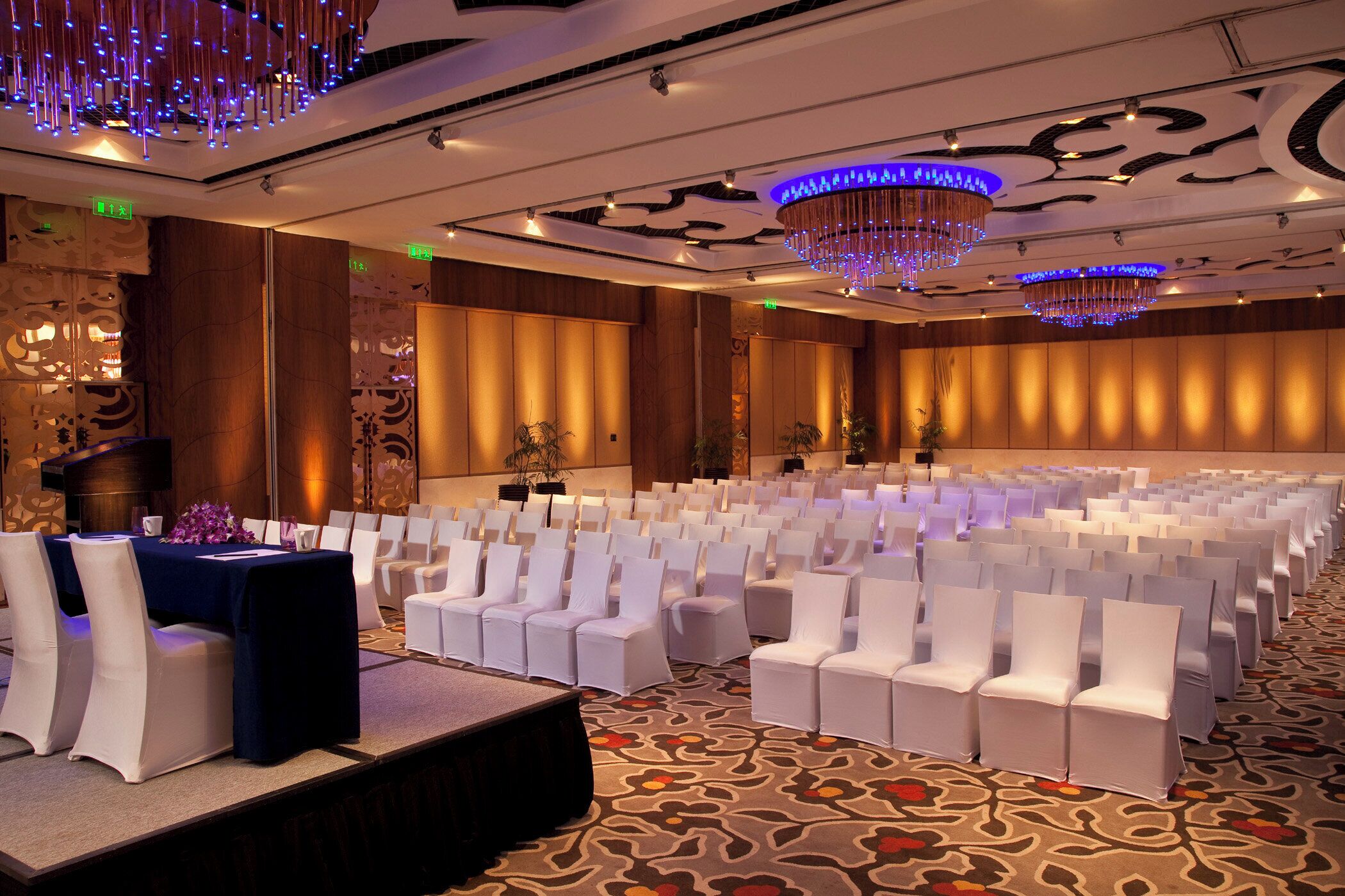 A conference room with rows of white chairs, a stage with a blue table, and decorative lighting.