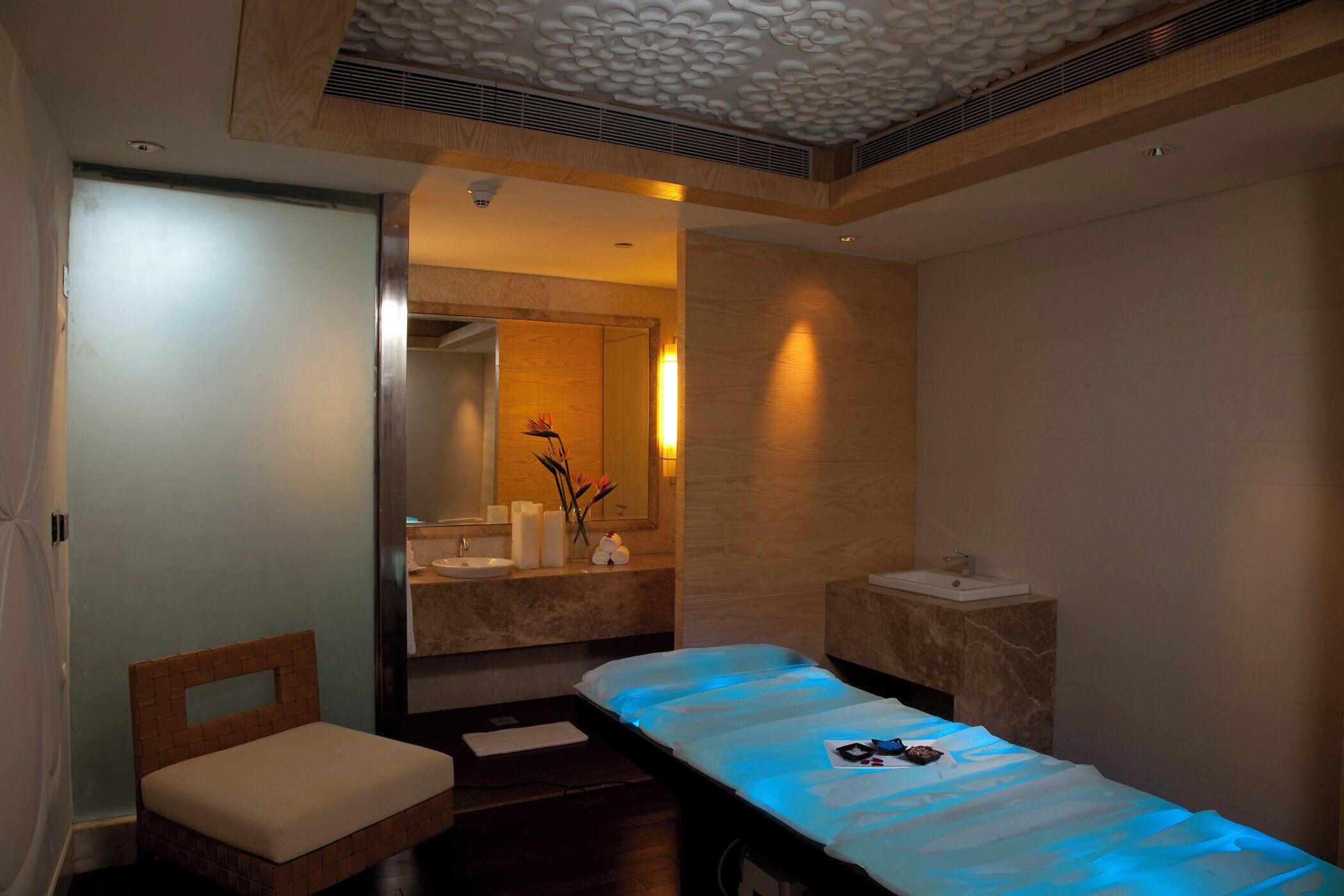 A modern spa room with a wooden bench, a large mirror, a sink, and a massage table with blue lighting.