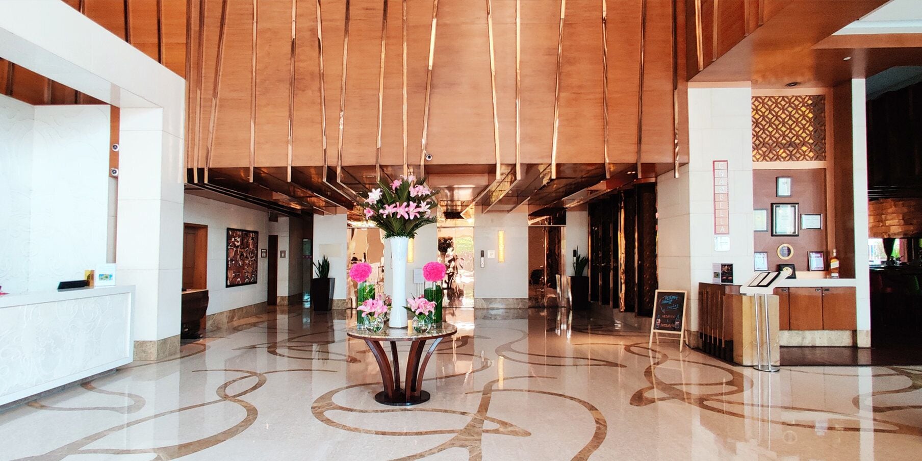 Un vestíbulo de hotel con un mostrador de recepción, un arreglo floral central y un diseño moderno de techo.