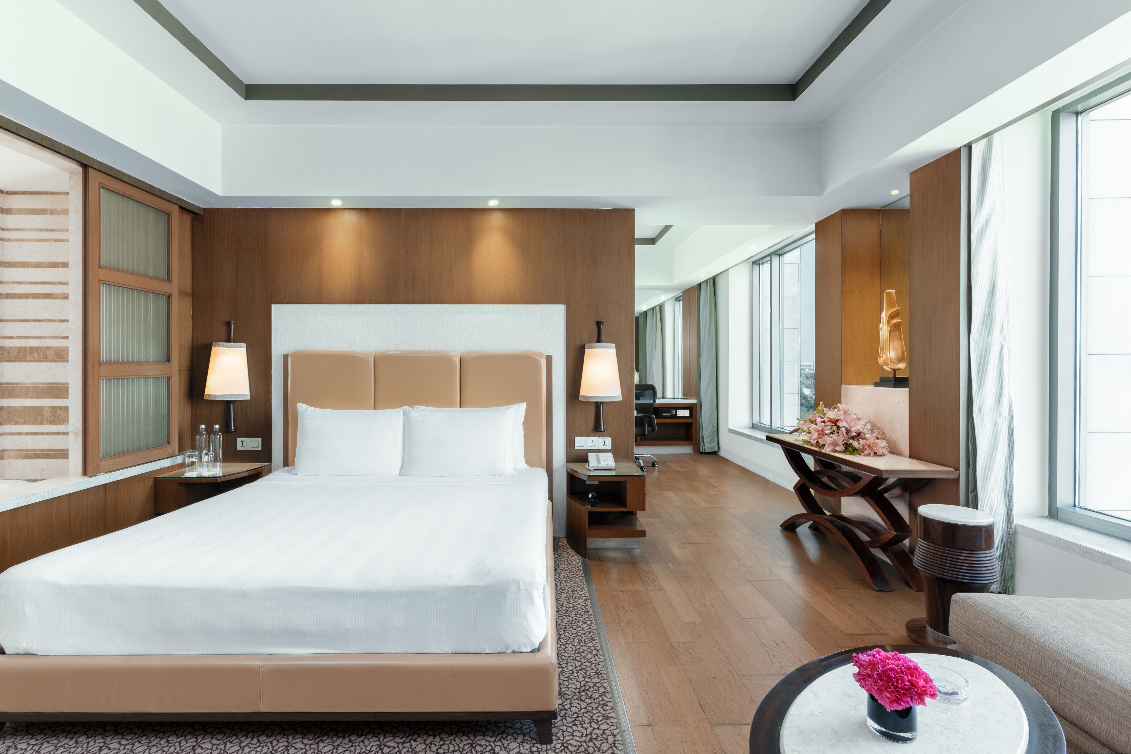 A modern hotel room with a large bed, bedside tables with lamps, a small desk, and a view of the city through large windows.