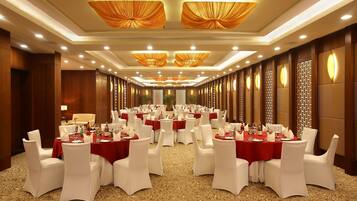 Uma sala de banquetes com mesas redondas, cadeiras brancas e toalhas vermelhas.