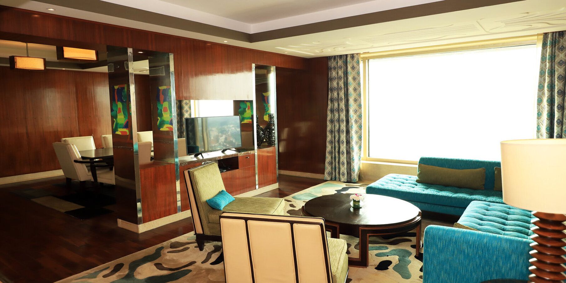 A hotel room with a dining area, a sofa, a TV, and a large window with curtains.