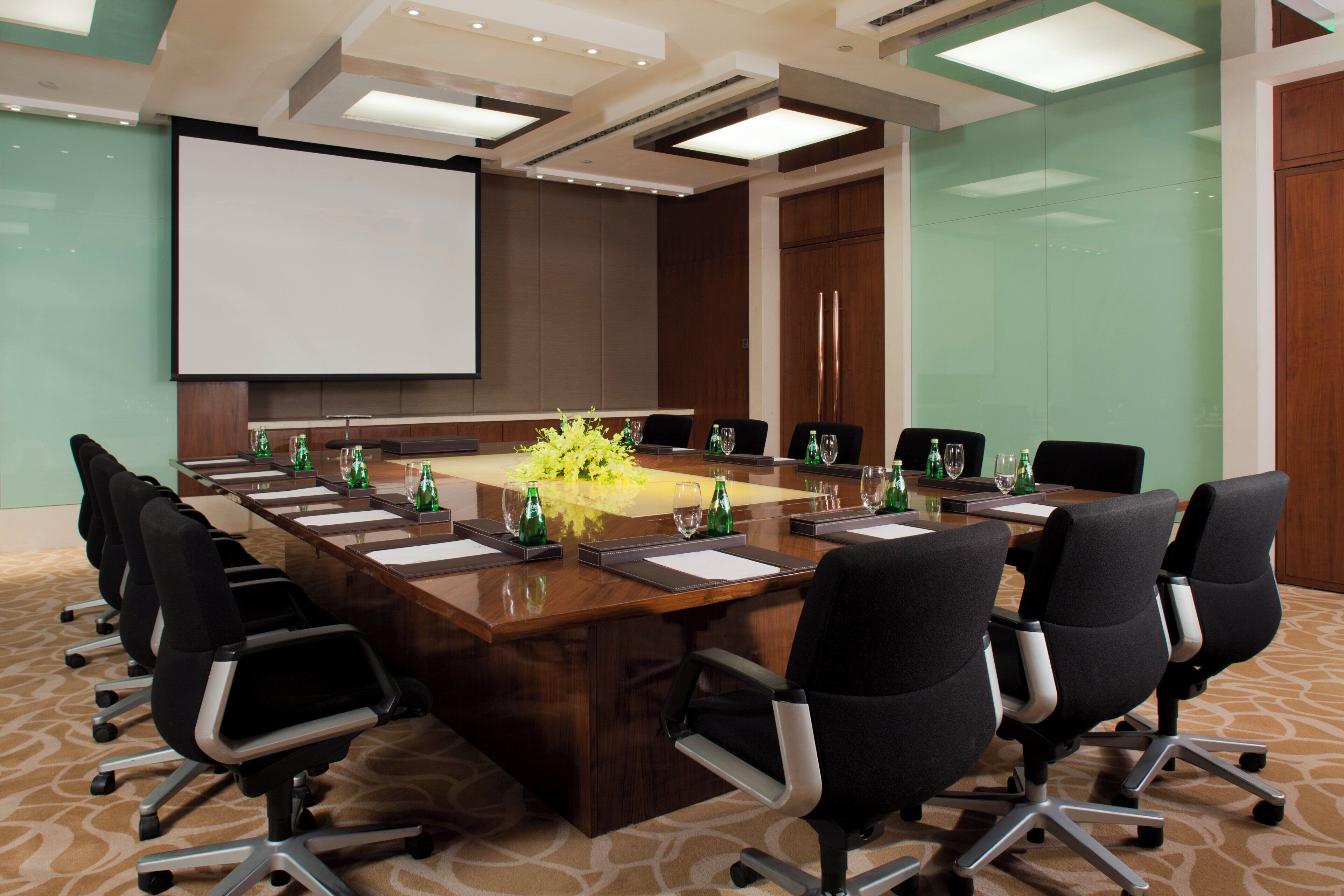 A conference room with a long table, chairs, a projector screen, and a floral centerpiece.
