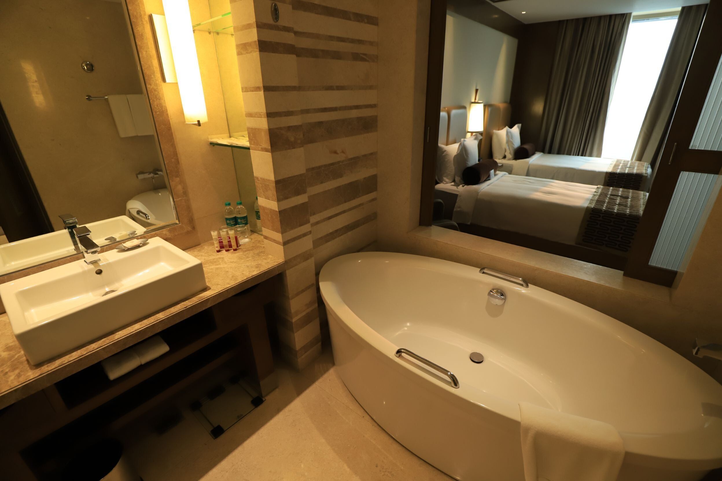 Une salle de bain d’hôtel avec deux lavabos, une baignoire et un grand miroir qui reflète une chambre à coucher avec un lit et des rideaux.