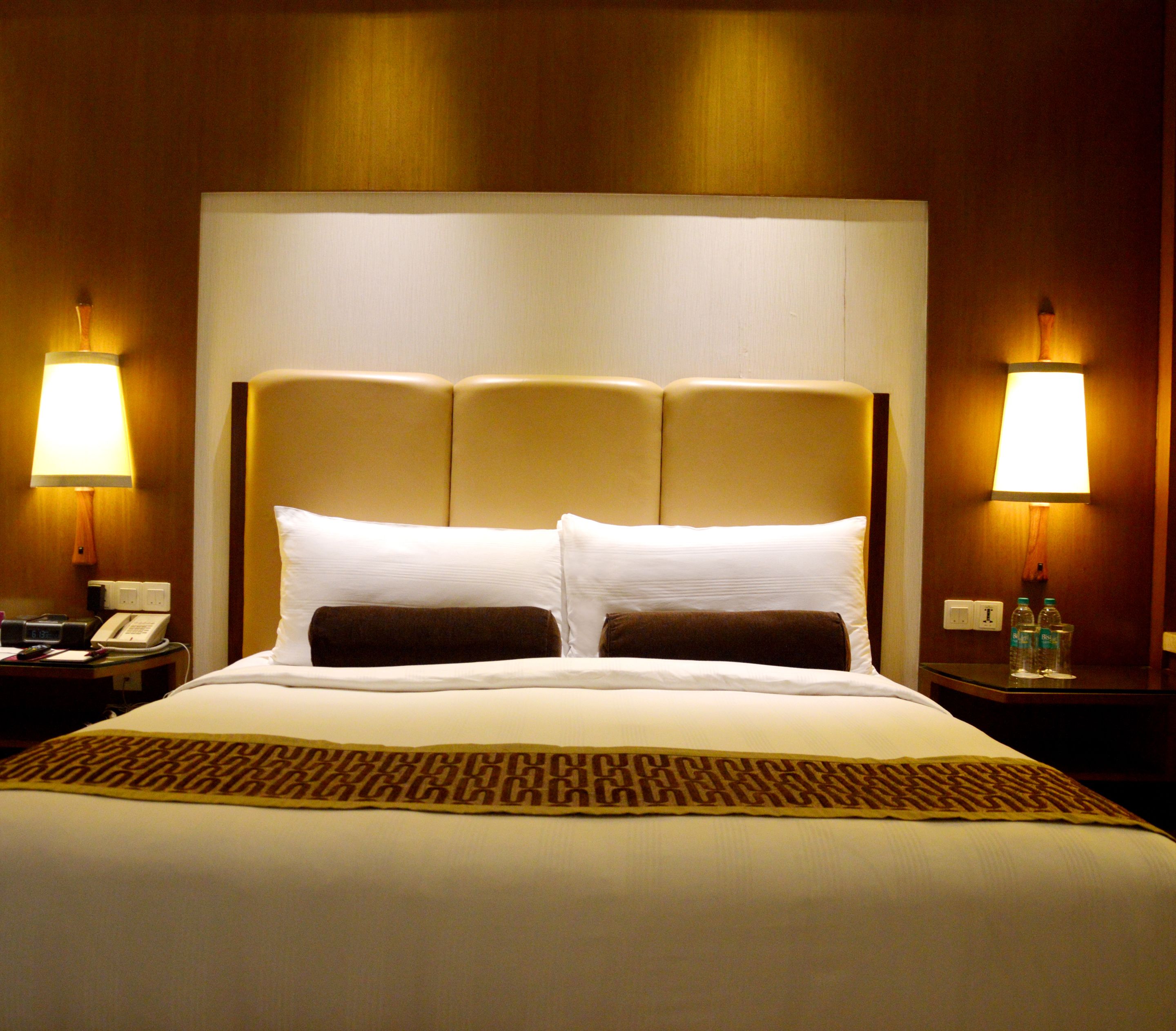 A hotel room with a large bed, two bedside lamps, a telephone, and a small table with a bottle and glasses.