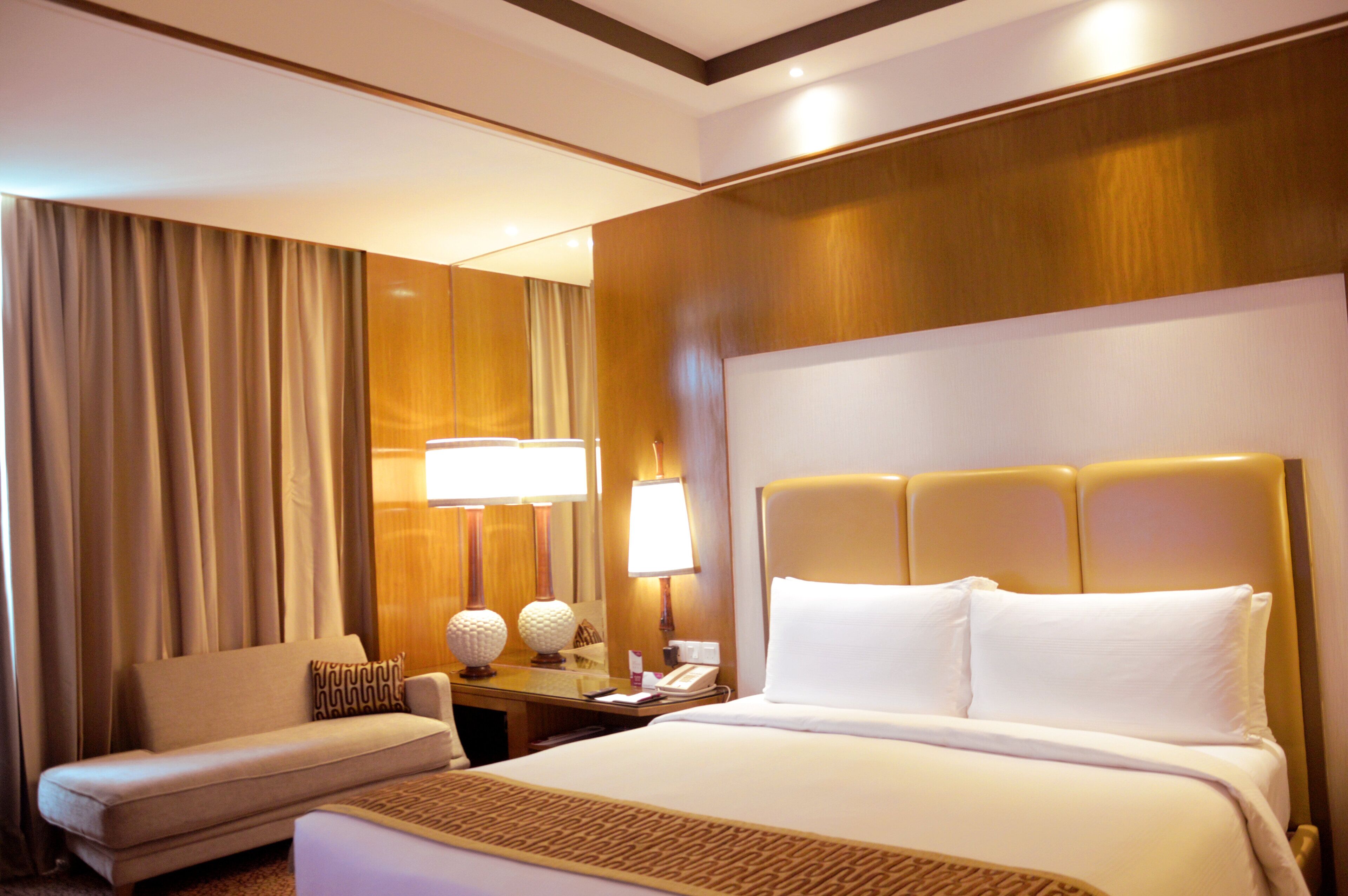 A hotel room with a bed, a sofa, a desk with lamps, and a large window with curtains.