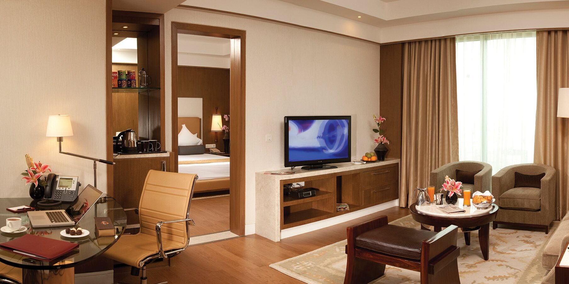 Ein Hotelzimmer mit einem Bett, einem Fernseher, einem Schreibtisch mit einem Laptop, einem Sessel, einem Couchtisch und einem Esstisch mit Frühstücksangeboten.