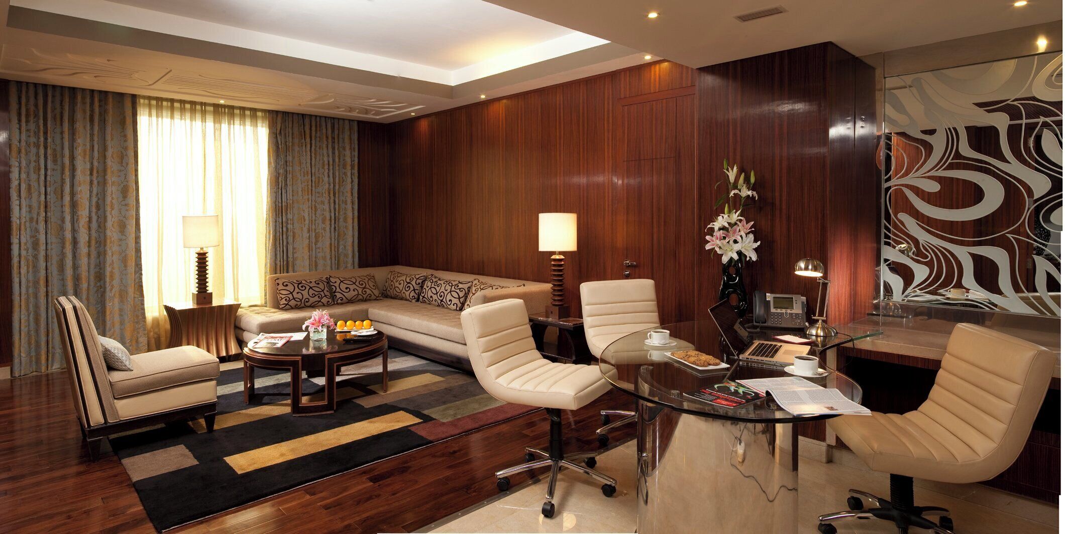A modern hotel room with a sofa, two armchairs, a coffee table, a desk with a computer, and a decorative wall piece.