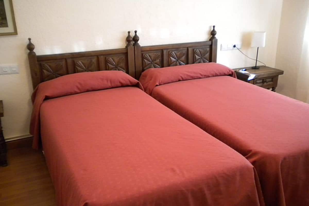 Una habitación con dos camas, cada una con cabecero de madera y ropa de cama roja. Hay una mesita de noche con una lámpara y un enchufe en la pared.