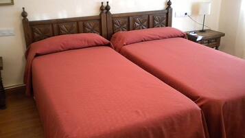 Una habitación con dos camas, cada una con cabecero de madera y ropa de cama roja. Hay una mesita de noche con una lámpara y un enchufe en la pared.
