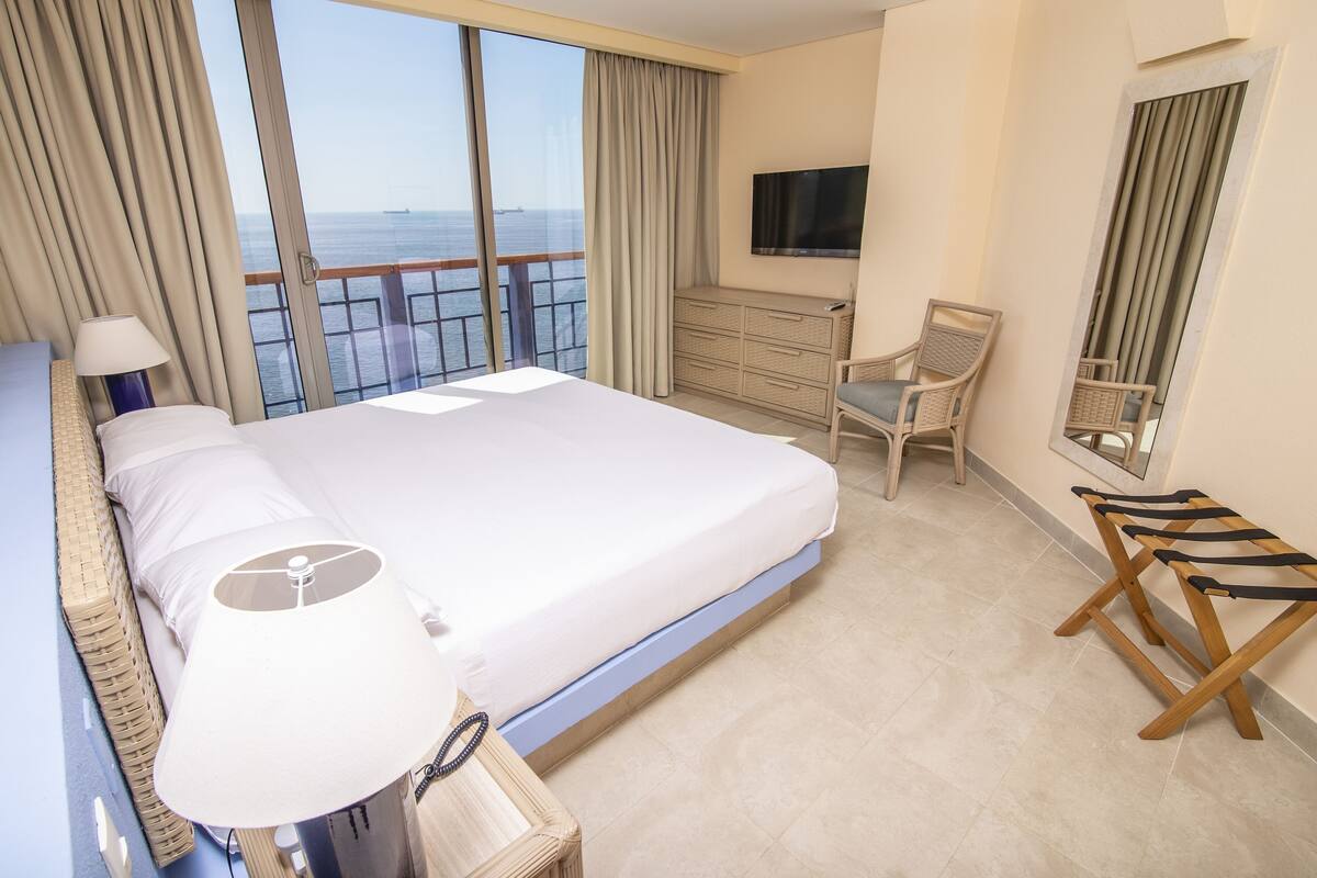 A hotel room with a bed, a TV, a desk, and a view of the sea.