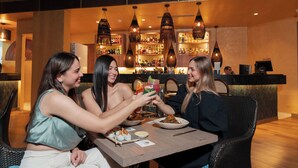 Tre personer sidder ved et bord med mad og drikke i en restaurant, hvor en bar ses i baggrunden.