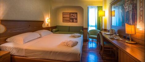 A hotel room with a large bed, a green sofa, a desk with a lamp, and a painting on the wall.