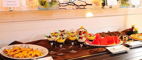 Una mesa de buffet con diversos alimentos y bebidas, como frutas, postres y un plato de fruta cortada.
