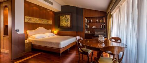 A hotel room with a bed, bedside table, lamp, wooden floor, dining table with chairs, and a bookshelf.