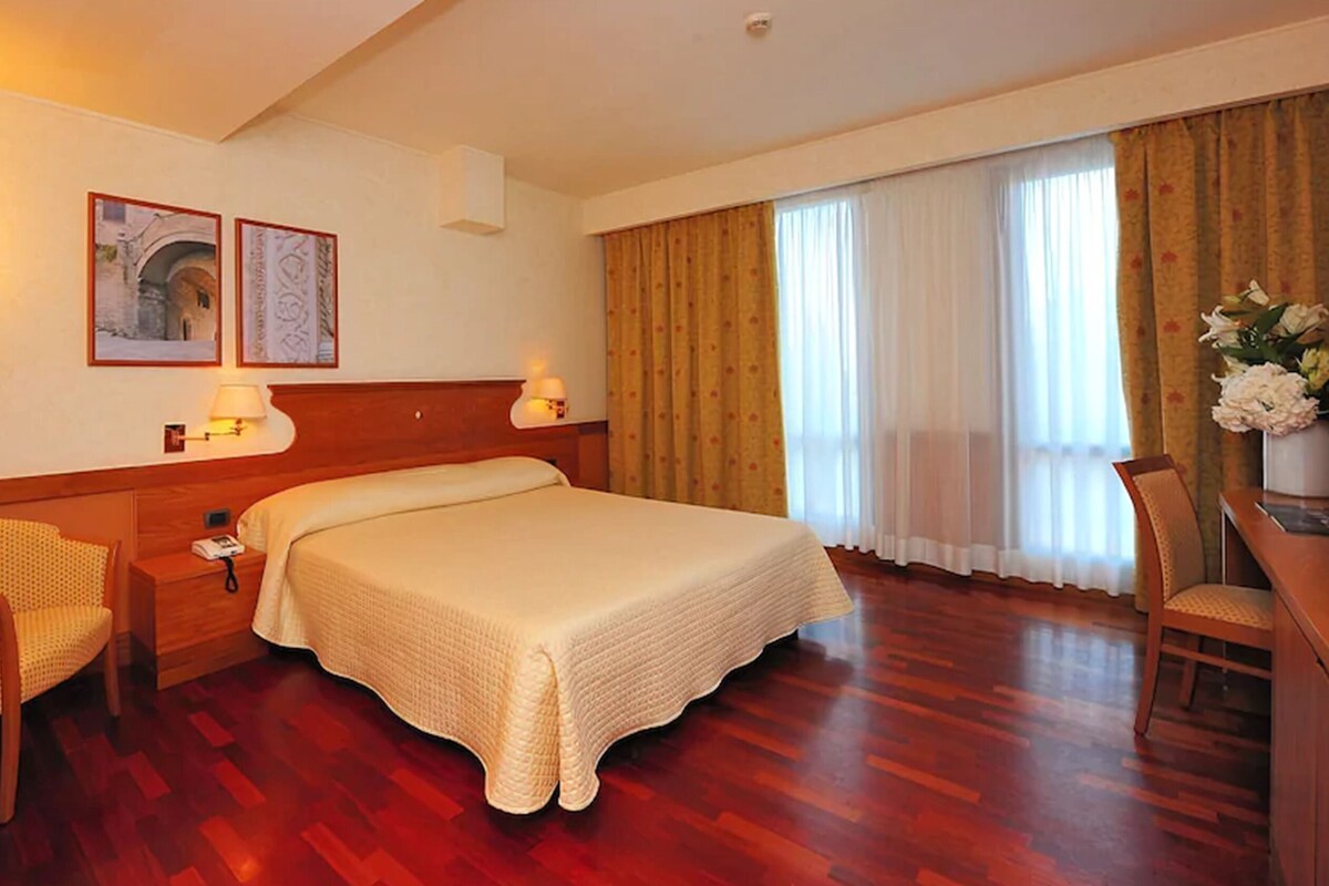 A hotel room with a large bed, two bedside tables with lamps, a chair, a desk with a vase of flowers, and a window with curtains.