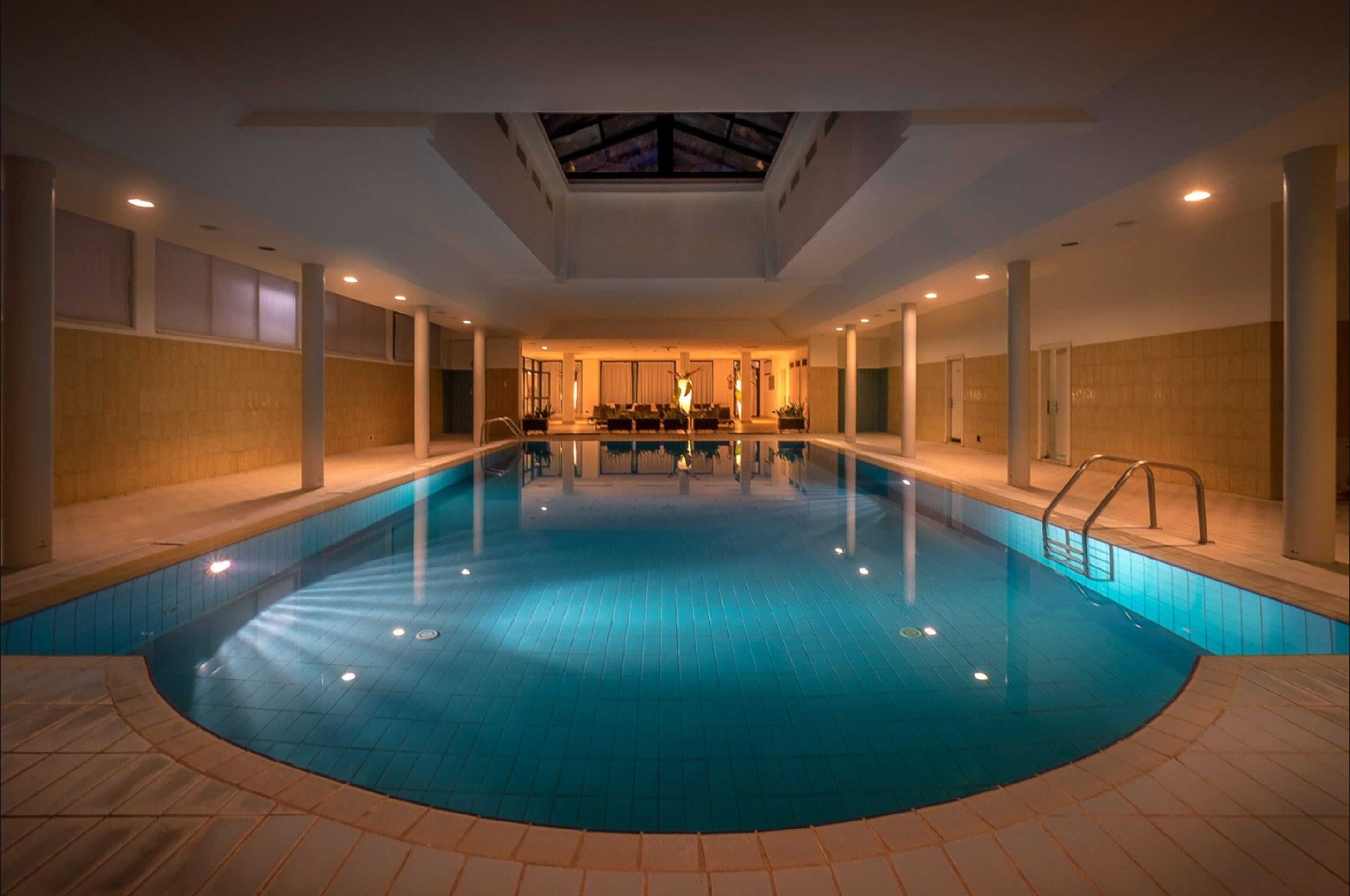 Una piscina interior con tragaluces, paredes de azulejos y una zona de estar al fondo.