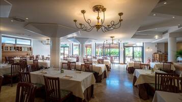 Un ristorante spazioso con numerosi tavoli e sedie, ampie finestre che lasciano entrare luce naturale e un lampadario decorativo.