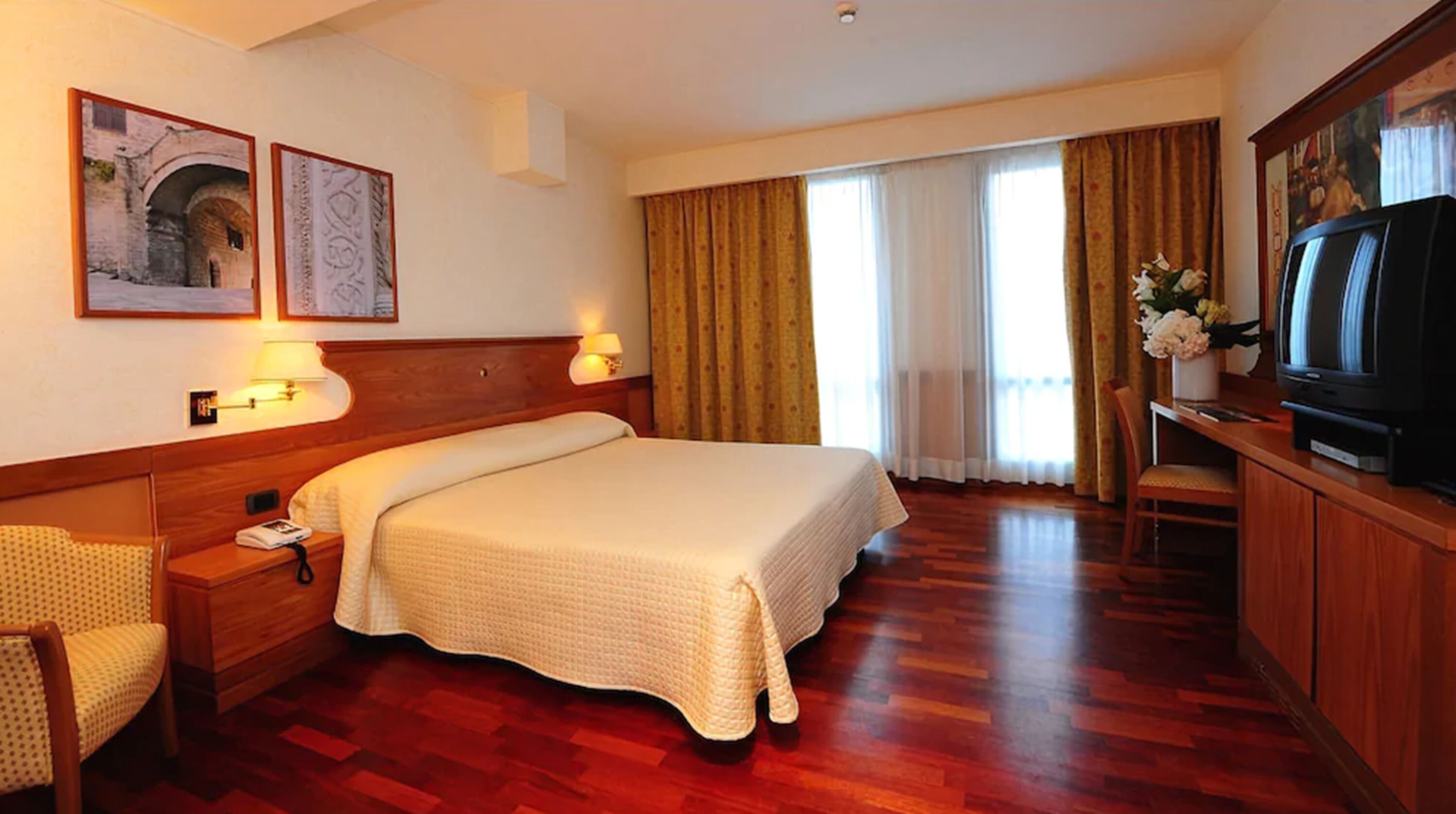 Habitación de hotel con una cama grande, cabecero de madera, dos lámparas fijadas a la pared, un televisor y un escritorio con una silla.