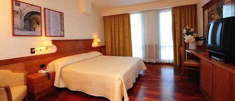 Habitación de hotel con una cama grande, cabecero de madera, dos lámparas fijadas a la pared, un televisor y un escritorio con una silla.