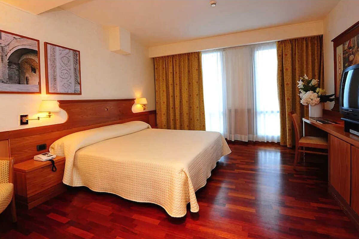 A hotel room with a large bed, wooden headboard, two wall-mounted lamps, a television, and a desk with a chair.