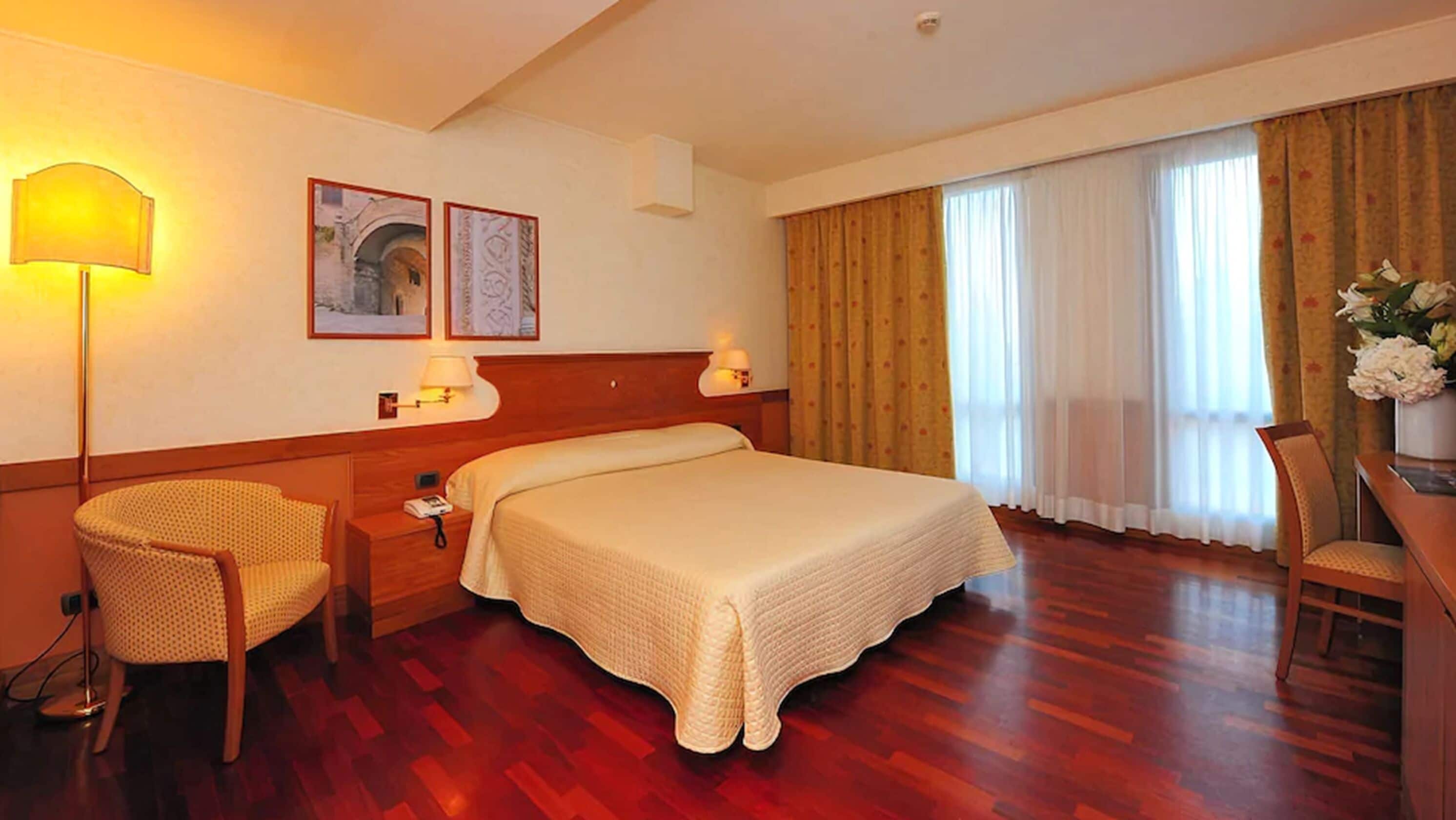 Une chambre d’hôtel avec un grand lit, deux lampes de chevet, une chaise, un bureau et une fenêtre avec des rideaux.