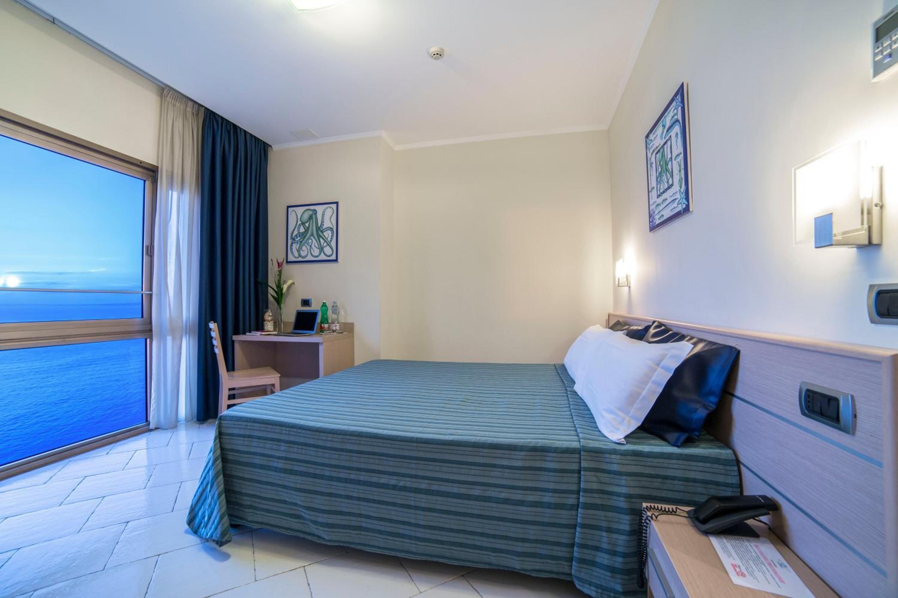 A hotel room with a bed, a desk, a chair, a nightstand, a telephone, a lamp, a picture frame, and a view of the sea.