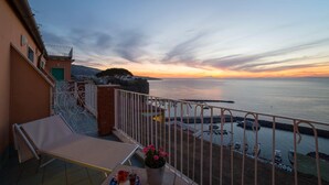 Un balcón con vista al mar al atardecer, una silla y una mesa con una maceta.