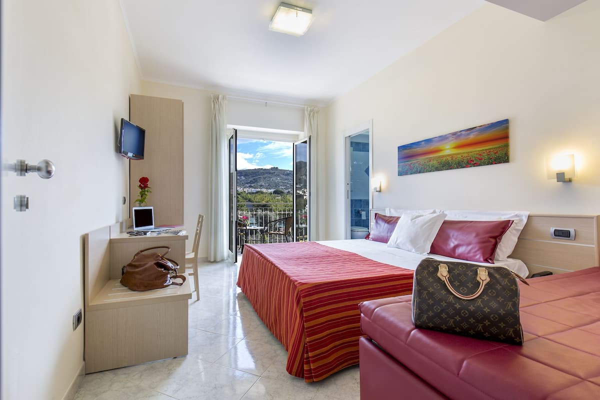 A hotel room with a bed, a desk, a chair, a red sofa, a TV, a painting, and a balcony with a view.