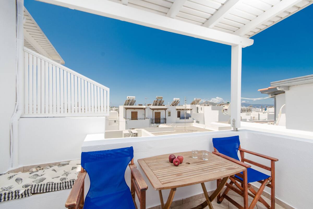 A balcony with a wooden table and two blue chairs, overlooking a row of white buildings.