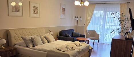 Habitación de hotel con una cama grande, una cama más pequeña, un escritorio y una silla. Hay dos mesitas de noche, una lámpara y una planta.