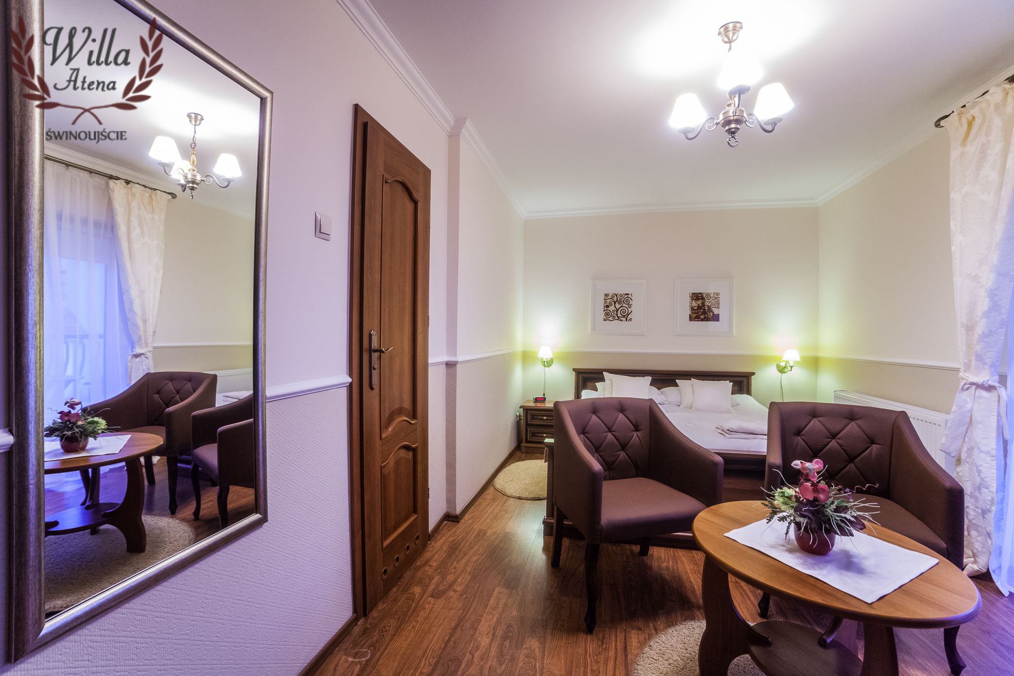 Ein Hotelzimmer mit Holzboden, einem Bett mit weißen Leinentüchern, zwei Polsterstühlen, einem runden Holztisch mit einer Vase, einem Spiegel und einer Kandelaber.