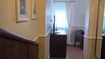A hallway with wooden flooring, a dark brown bench, and a small table with a television. There are framed pictures on the wall and a window with white curtains.