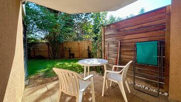 Un patio con una mesa redonda y dos sillas, un cerco de madera y un estante metálico con un objeto verde.
