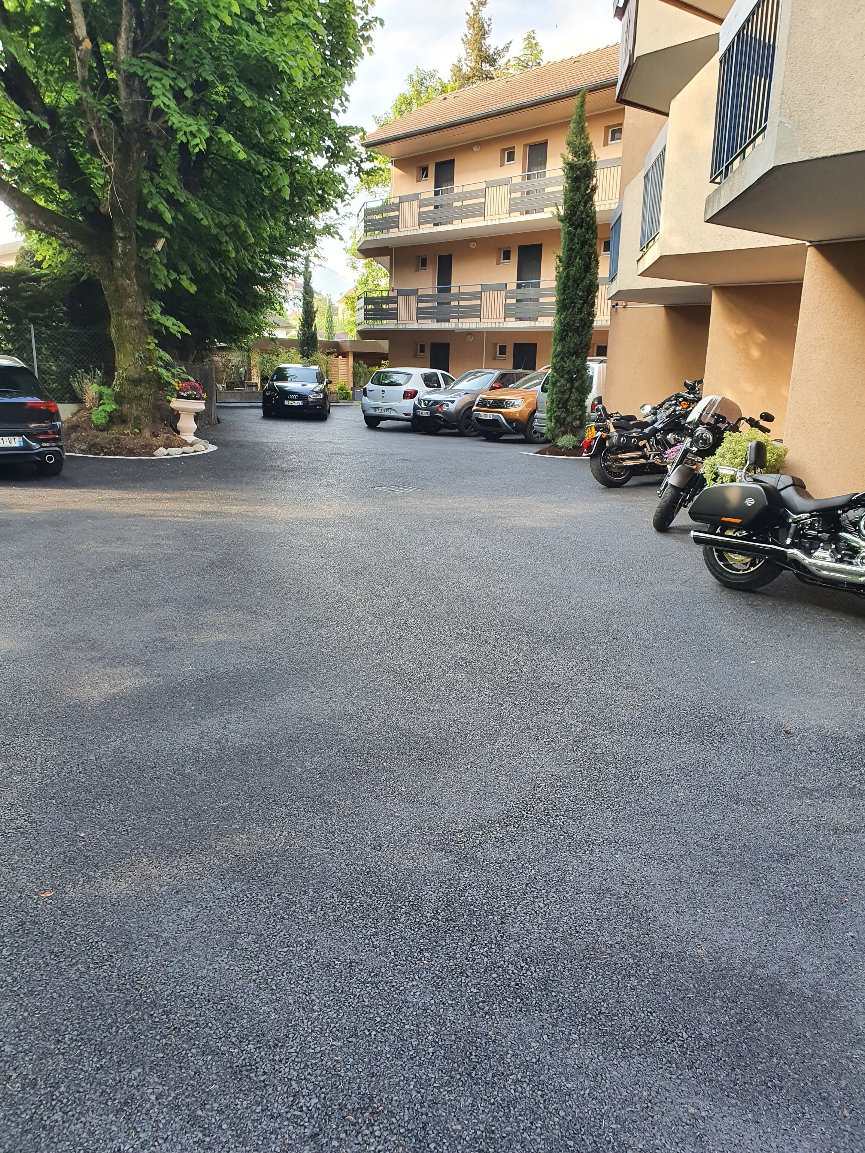 A parking area with several cars and motorcycles, a tree, and a building with balconies.