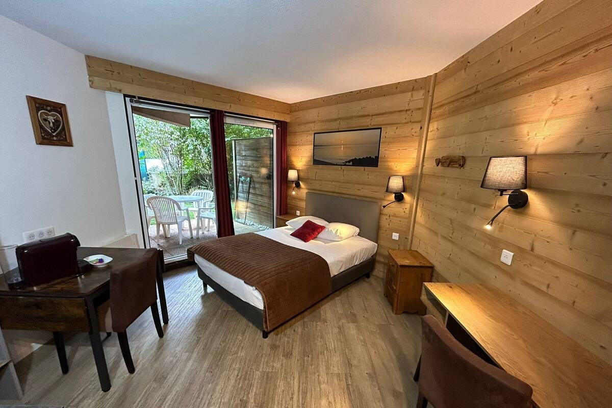 A wooden-paneled room with a bed, a desk, and a chair. There is a view through a glass door to an outdoor area with chairs.