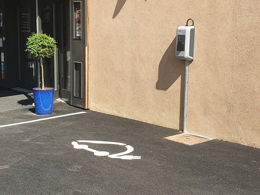 Ein Parkplatz mit Rollstuhlsymbol, ein blauer Pflanztopf mit einem kleinen Baum und eine Überwachungskamera an einer beigen Wand.